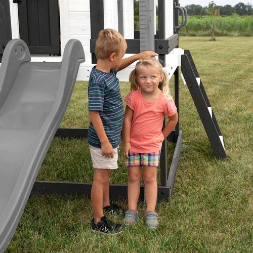 Casavibeo Backyard Discovery Sweetwater Heights elevated playhouse with a cozy porch and slide, set in a lush backyard.