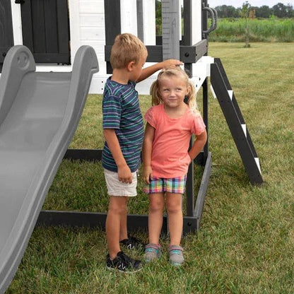 Casavibeo Backyard Discovery Sweetwater Heights elevated playhouse with a cozy porch and slide, set in a lush backyard.